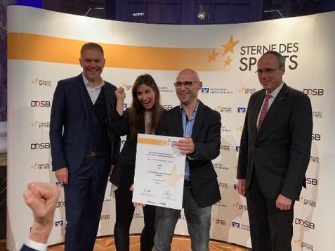 Vier Personen stehen an einem Hintergrund mit dem Schriftzug „Sterne des Sports“ und präsentieren stolz eine Urkunde.