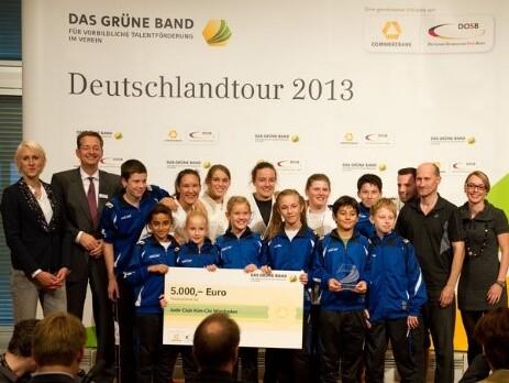 Gruppenszene mit Jugendlichen in Sportkleidung, die einen Scheck &uuml;ber 5.000 Euro halten, vor einer Pr&auml;sentationswand.