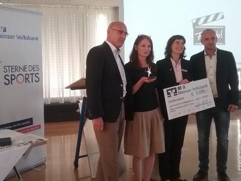 Vier Personen stehen auf einer Veranstaltung, halten einen Scheck &uuml;ber 5.000 Euro und stehen neben einem Banner der Mainzer Volksbank.