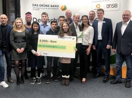 Gruppenzusammenkunft mit Jugendlichen und Erwachsenen, die einen Scheck &uuml;ber 5.000 Euro vor einem Banner pr&auml;sentieren.