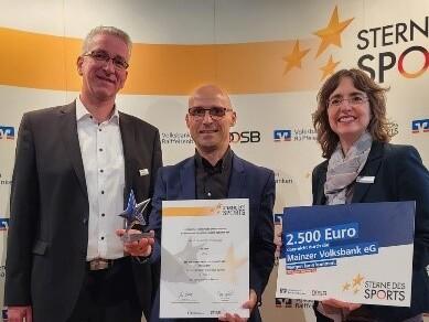 Drei Personen halten eine Auszeichnung und einen Scheck &uuml;ber 2.500 Euro vor einem Banner mit "Sterne des Sports".