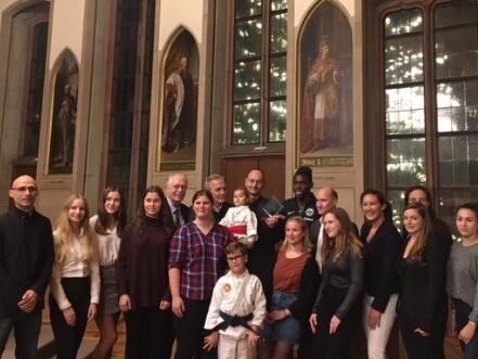 Gruppierung von mehreren Personen vor historischen Fenstern in einer Halle, mit Gem&auml;lden im Hintergrund.
