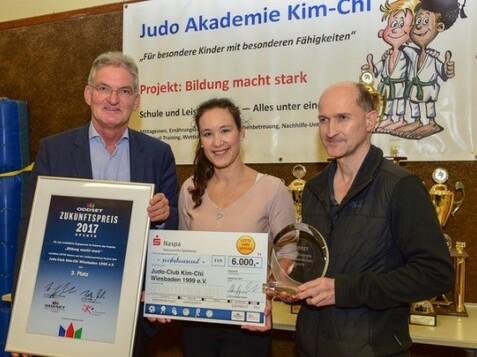 Drei Personen stehen l&auml;chelnd vor einem Banner und halten Auszeichnungen der Judo Akademie Kim-Chi in der Hand.