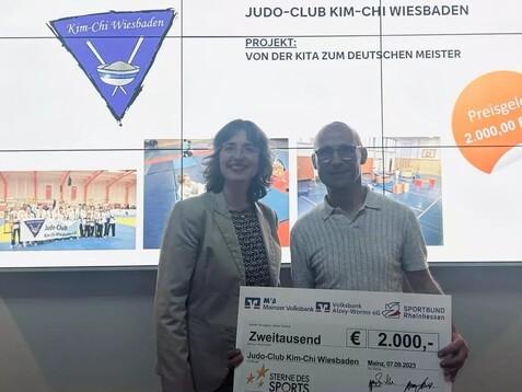 Frau und Mann halten einen Scheck &uuml;ber 2000 Euro f&uuml;r den Judo-Club Kim-Chi Wiesbaden bei einer Preisverleihung.