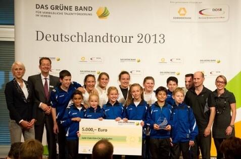 Gruppenszene mit Jugendlichen in Sportkleidung, die einen Scheck &uuml;ber 5.000 Euro halten, vor einer Pr&auml;sentationswand.