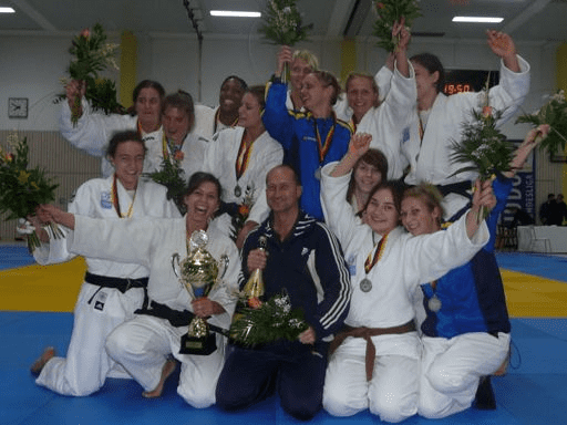 Judo-Team feiert Sieg mit Trophäen und Blumen auf der Matte in einer Halle, umgeben von jubelnden Spielern.