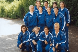 Gruppe von Mädchen und Frauen in blauen Sportanzügen lächelt auf einem gepflasterten Platz vor grüner Kulisse.