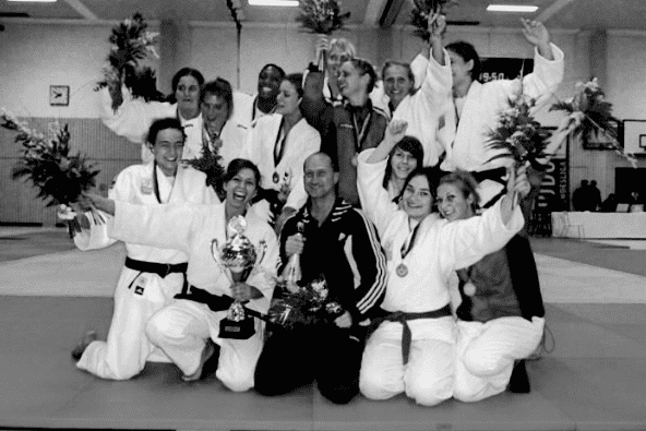 Gruppennahaufnahme von Judokas mit Pokalen und Blumen, die jubeln und Medaillen tragen, auf einer Judomatte.