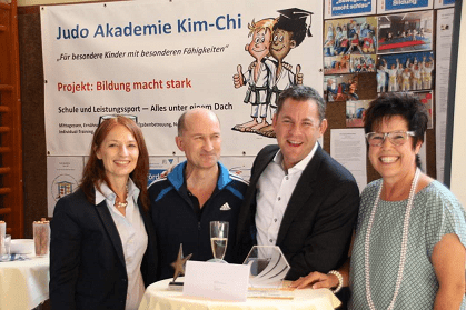 Vier Personen stehen lächelnd an einem Stehtisch, im Hintergrund ein Plakat der Judo Akademie Kim-Chi über Bildung.