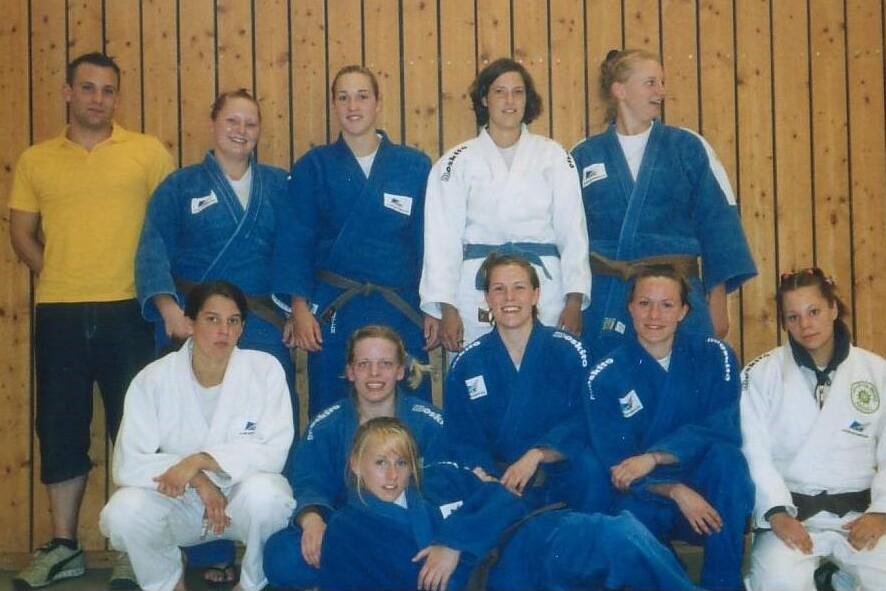 Gruppenausstellung von Judo-Kämpfern in blauen und weißen Anzügen, aufgestellt vor einer Holzwand.