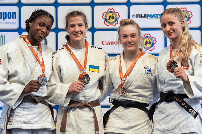 Vier Judoka in weißen Anzügen mit Medaillen posieren gemeinsam vor einer Sponsorenwand.