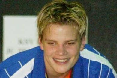 Lächelnder junger Sportler in blauer Jacke hält eine Goldmedaille und eine gelbe Rose in der Hand.