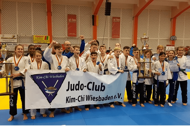 Gruppenshot des Judo-Teams Kim-Chi Wiesbaden mit Medaillen, Pokalen und Banner auf einer blauen Matte.