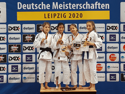Vier junge Judoka in weißen Gis stehen auf dem Podium, halten Medaillen und Urkunden, vor einer Wand mit Sponsorenlogos.