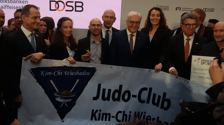 Gruppenszene mit Menschen, die ein Banner des Judo-Clubs Kim-Chi Wiesbaden halten, während sie an einer Veranstaltung teilnehmen.