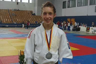 Junger Judoka in Weiß mit Silbermedaille, Blumenstrauß und Urkunde steht auf einer Judomatte.