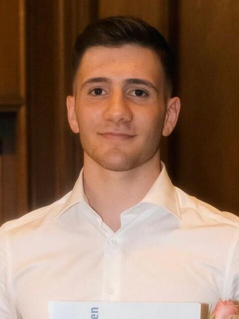 Junger Mann in wei&szlig;em Hemd h&auml;lt ein Abschlusszeugnis und eine Rose in der Hand, vor einem Holzpaneel.