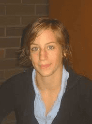 Portr&auml;t einer Person mit kurzen Haaren, die einen schwarzen Pullover und ein blaues Hemd tr&auml;gt, vor einer Ziegelwand.