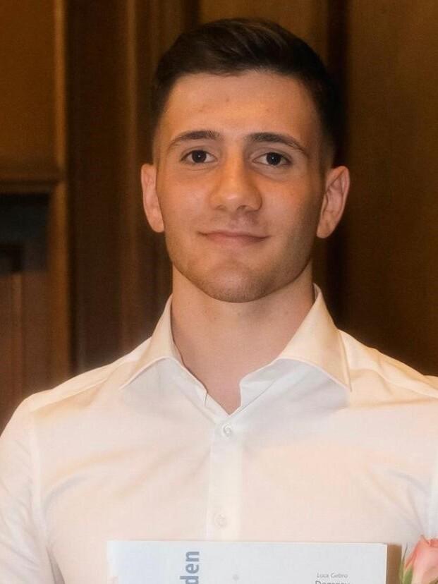 Junger Mann in weißem Hemd hält ein Abschlusszeugnis und eine Rose in der Hand, vor einem Holzpaneel.