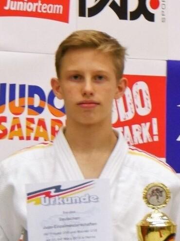Junger Judoka in wei&szlig;em Gi h&auml;lt eine Urkunde und einen Pokal, vor einem Hintergrund mit Sportwerbung.