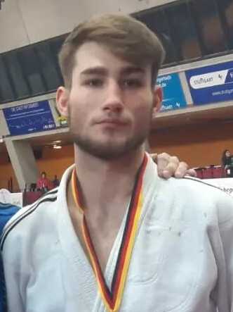 Mann im Judoanzug mit Medaille steht neben einem Trainer auf einer Judo-Matte in einer Sporthalle.