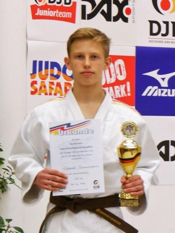Junger Judoka in weißem Gi hält eine Urkunde und einen Pokal, vor einem Hintergrund mit Sportwerbung.