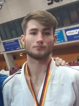 Ein junger Judoka in wei&szlig;em Gi mit Medaille steht neben einem Trainer in blauer Jacke auf einer Judomatte.