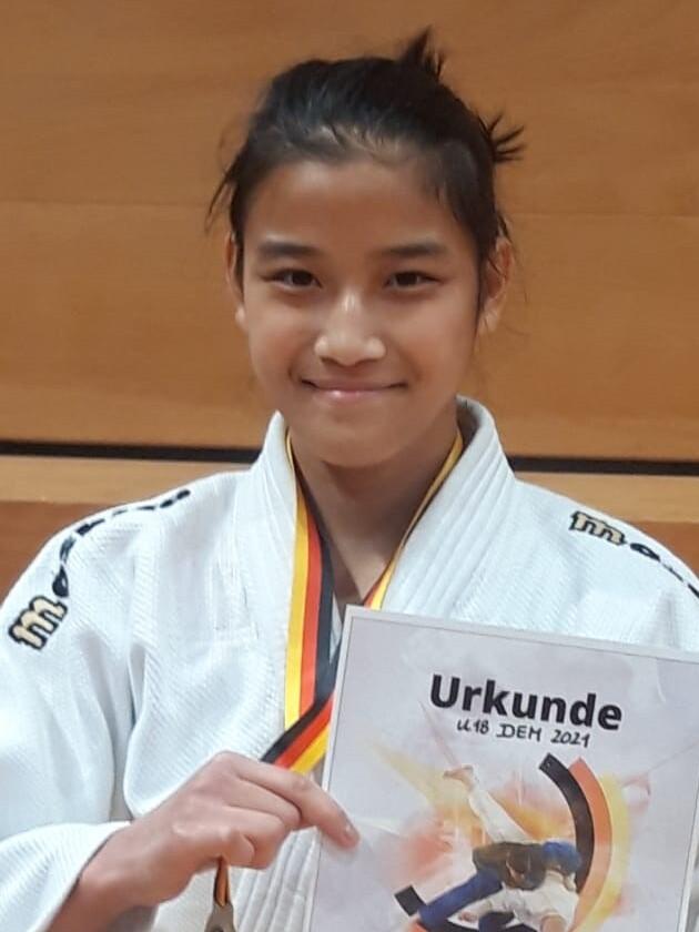 Junge in Judogi h&auml;lt Urkunde und Medaille nach Wettkampf, mit Siegerpose vor Holztrib&uuml;ne.