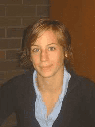 Portr&auml;t einer Person mit kurzen Haaren, die einen schwarzen Pullover und ein blaues Hemd tr&auml;gt, vor einer Ziegelwand.