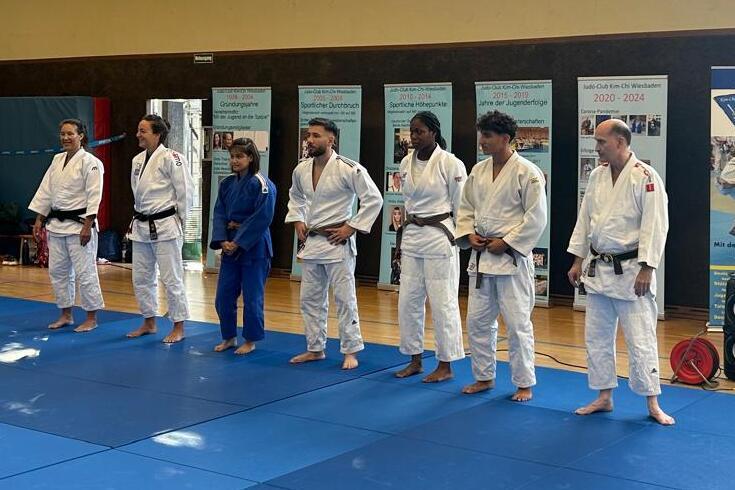 Judo-Training in einer Halle: sechs Judoka in Uniform stehen auf einer blauen Matte, Zuschauer im Hintergrund.