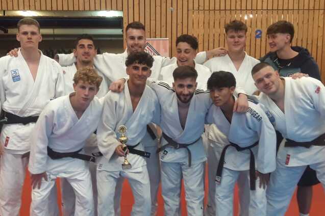 Gruppe von zehn Judokas in wei&szlig;en Anz&uuml;gen posiert gemeinsam stolz mit einer Troph&auml;e auf einer Judomatte.