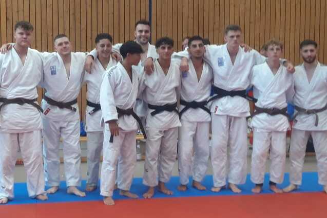 Gruppe von zehn Judokas in wei&szlig;en und schwarzen Gis, die l&auml;chelnd auf einer Judomatte posieren.