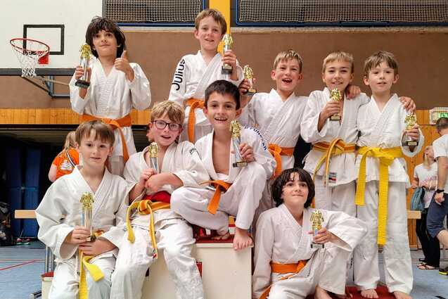 Gruppe von zehn Kindern in wei&szlig;en Judokimonos mit orangefarbenen G&uuml;rteln, auf einer Siegertreppe mit Pokalen.