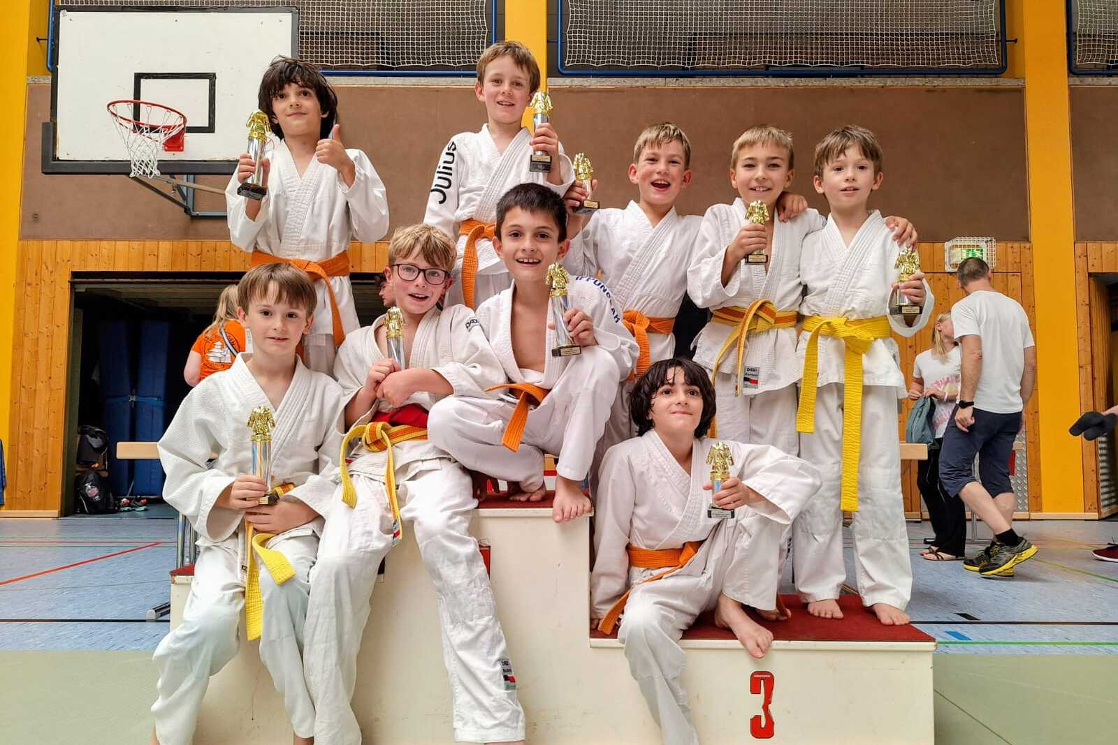 Gruppe von zehn Kindern in weißen Judokimonos mit orangefarbenen Gürteln, auf einer Siegertreppe mit Pokalen.