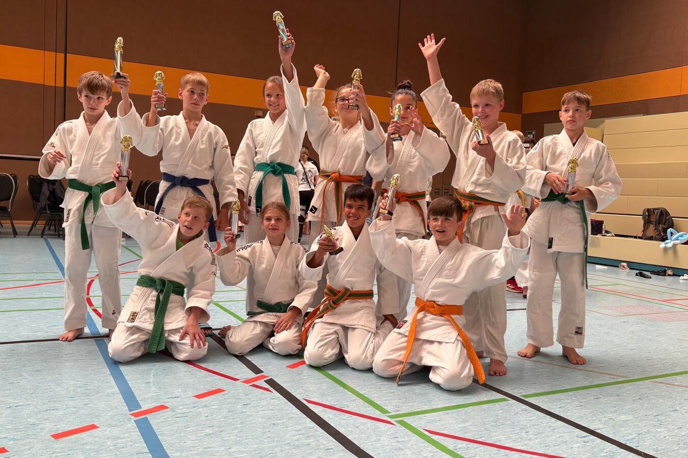 Gruppe von Kindern in Karateanzügen mit Medaillen in einer Turnhalle, einige heben die Arme triumphierend.