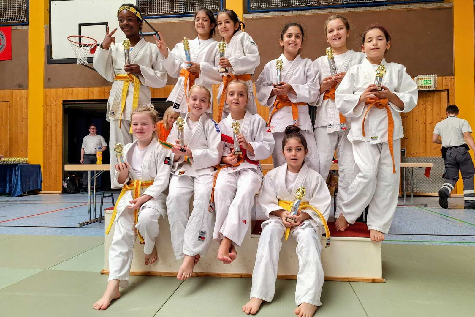 Gruppe von zehn Mädchen in Karateanzügen mit Medaillen, die auf einer Plattform stehen, im Hintergrund eine Sporthalle.