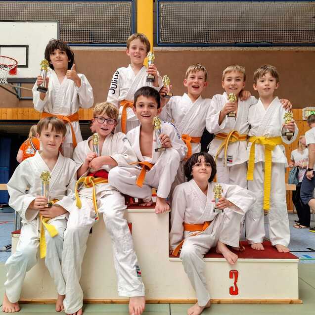 Gruppe von zehn Kindern in wei&szlig;en Judokimonos mit orangefarbenen G&uuml;rteln, auf einer Siegertreppe mit Pokalen.