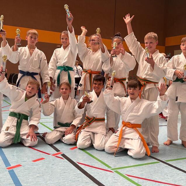 Gruppe von Kindern in Karateanz&uuml;gen mit Medaillen in einer Turnhalle, einige heben die Arme triumphierend.