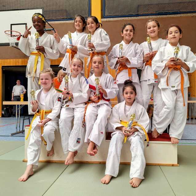 Gruppe von zehn M&auml;dchen in Karateanz&uuml;gen mit Medaillen, die auf einer Plattform stehen, im Hintergrund eine Sporthalle.