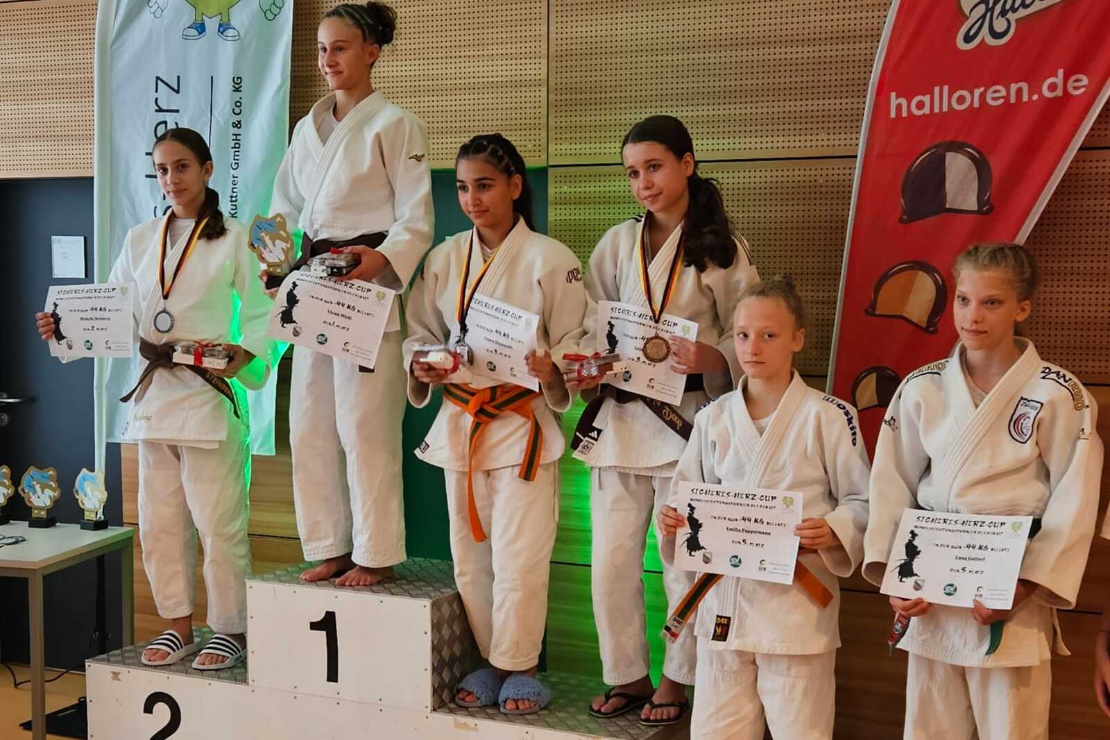 Sechs Judoka in weißem Gi stehen auf einem Siegerpodest, halten Urkunden und Medaillen nach einem Wettkampf.