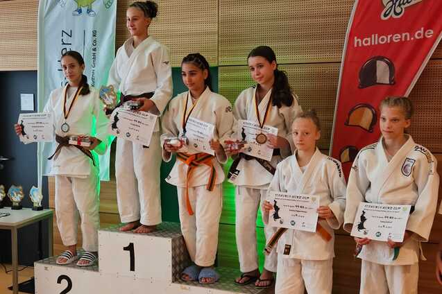 Sechs Judoka in wei&szlig;em Gi stehen auf einem Siegerpodest, halten Urkunden und Medaillen nach einem Wettkampf.