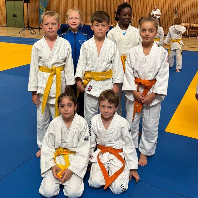 Sechs Kinder in Judogis stehen auf einer blauen Judomatte, einige tragen gelbe und orange G&uuml;rteln.