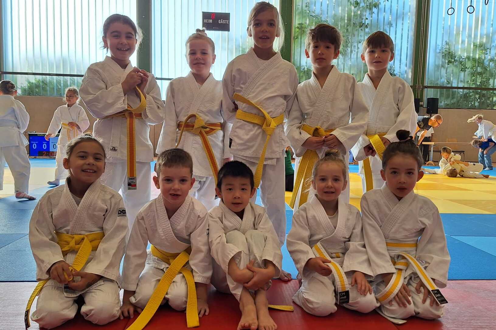 Gruppe von zehn Kindern in wei&szlig;en Karateanz&uuml;gen mit gelben G&uuml;rteln, stehend und sitzend auf einer Trainingsfl&auml;che.