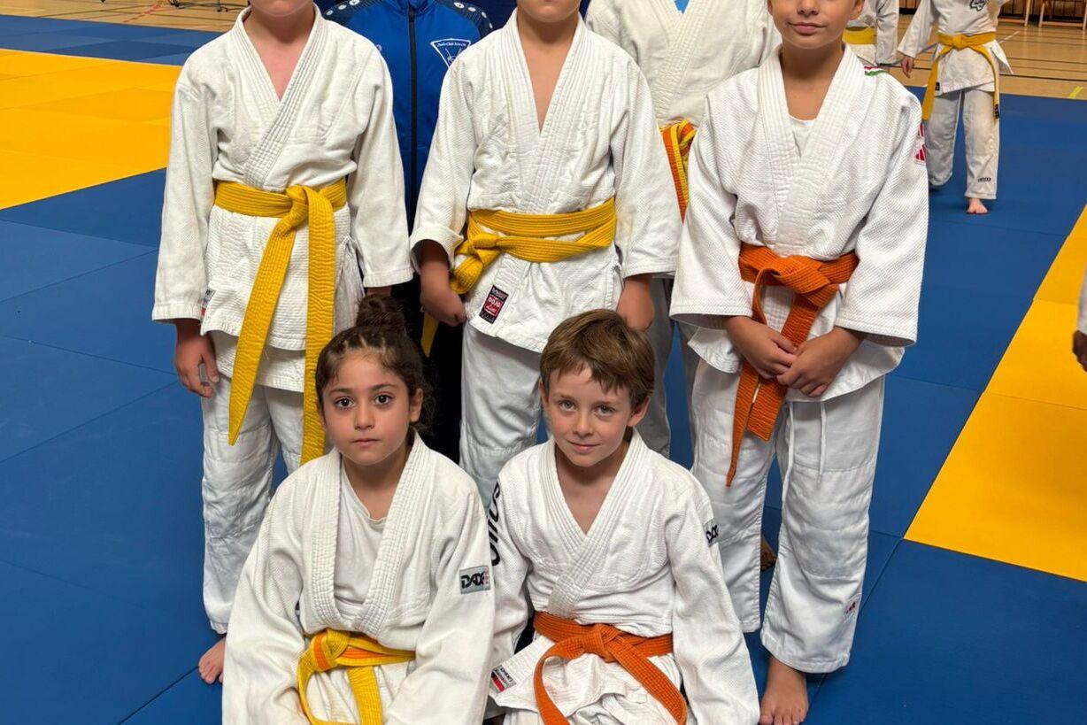 Sechs Kinder in Judogis stehen auf einer blauen Judomatte, einige tragen gelbe und orange G&uuml;rteln.