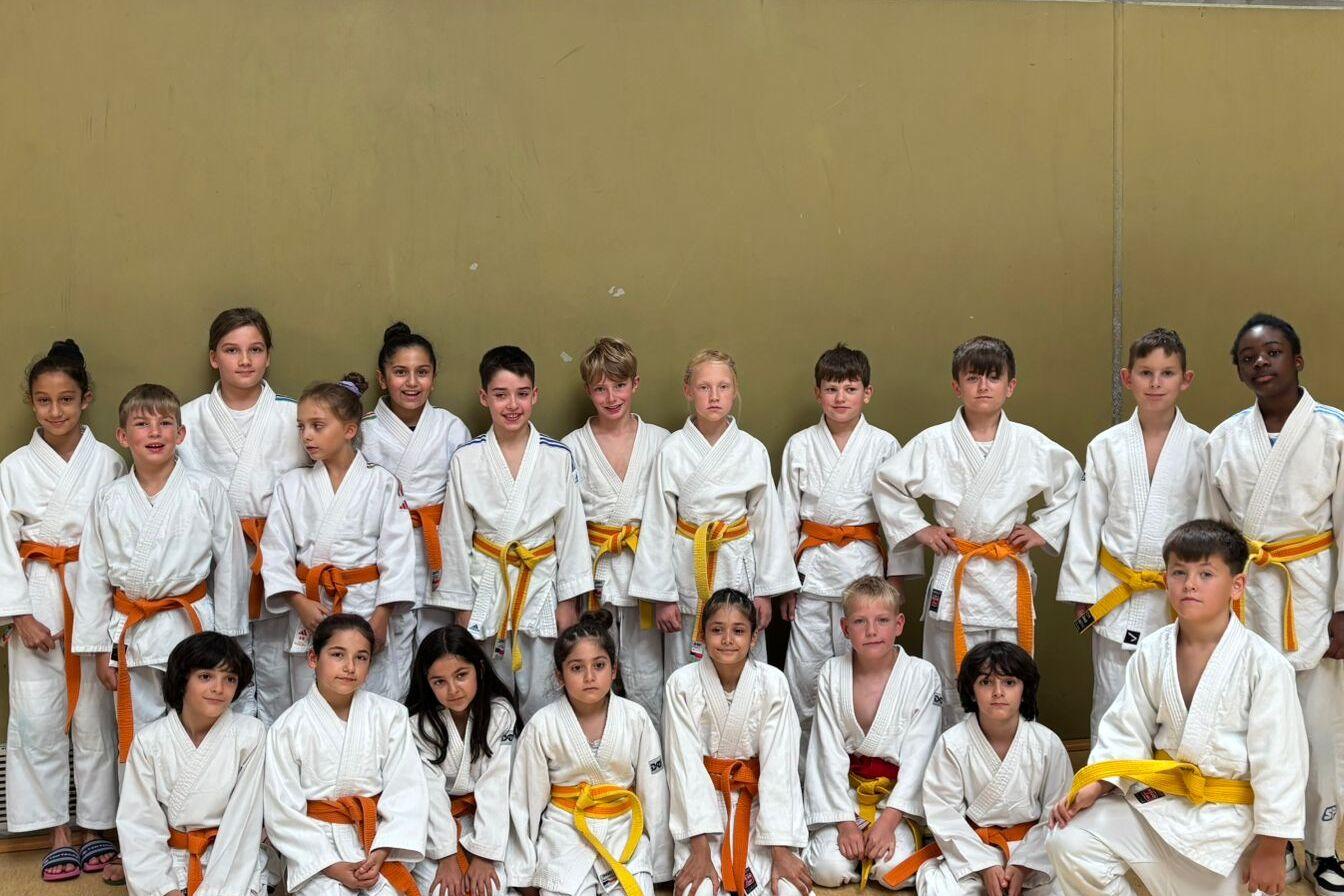 Gruppe von Karate-Kindern in wei&szlig;en Anz&uuml;gen mit orangefarbenen G&uuml;rteln, versammelt in einer Sporthalle.