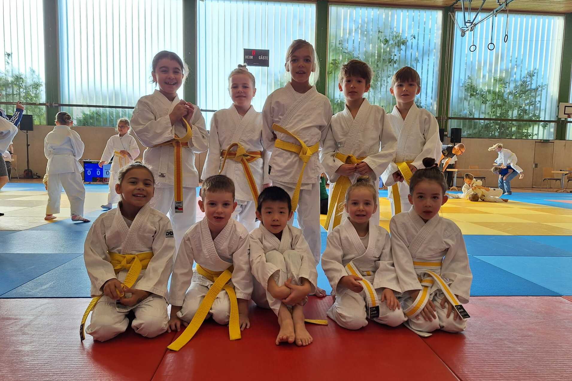 Gruppe von zehn Kindern in wei&szlig;en Karateanz&uuml;gen mit gelben G&uuml;rteln, stehend und sitzend auf einer Trainingsfl&auml;che.