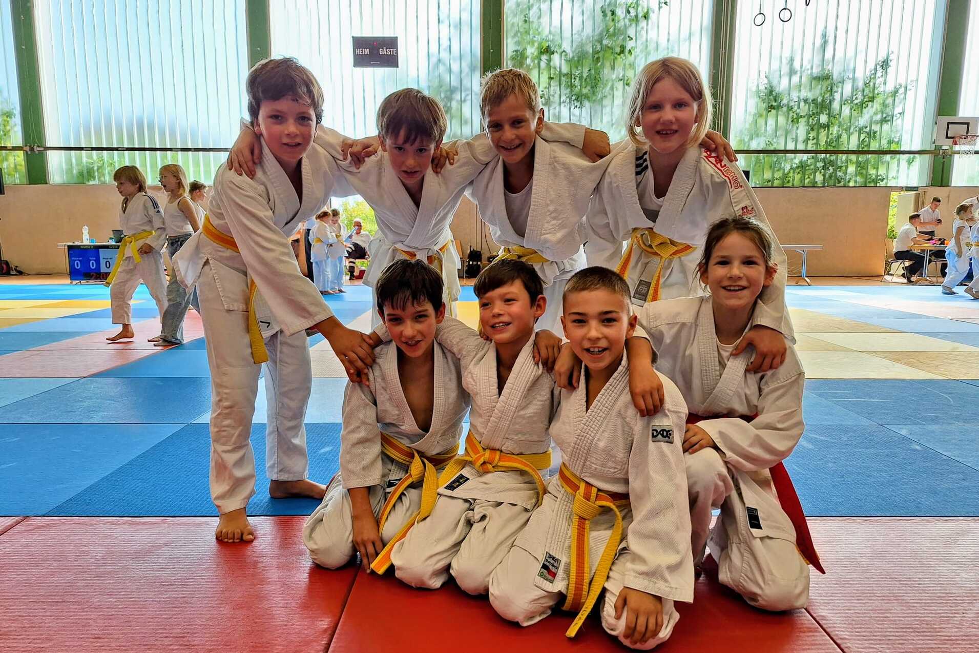 Gruppe von acht Kindern in wei&szlig;en Judokost&uuml;men mit gelben G&uuml;rteln posiert l&auml;chelnd auf einer Judomatte.