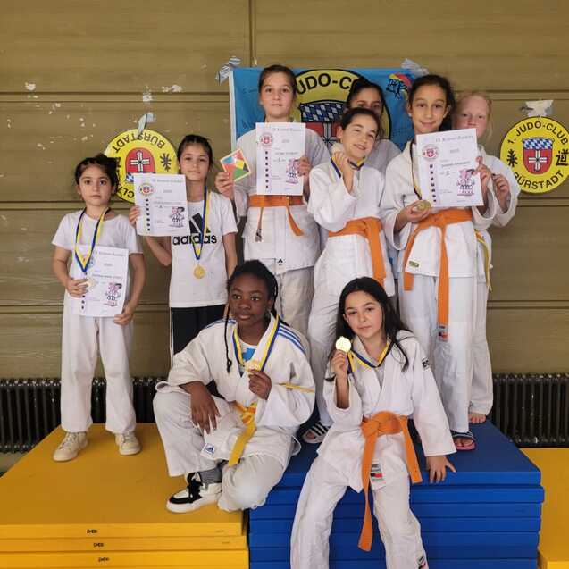 Gruppenfoto von sieben jungen Judokas in wei&szlig;en Anz&uuml;gen mit Medaillen und Urkunden auf bunten Wettkampfb&ouml;den.