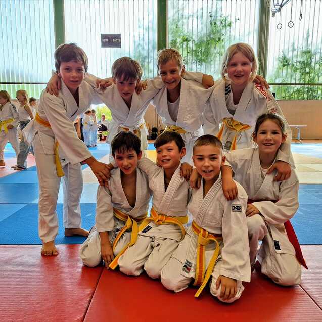 Gruppe von acht Kindern in wei&szlig;en Judokost&uuml;men mit gelben G&uuml;rteln posiert l&auml;chelnd auf einer Judomatte.