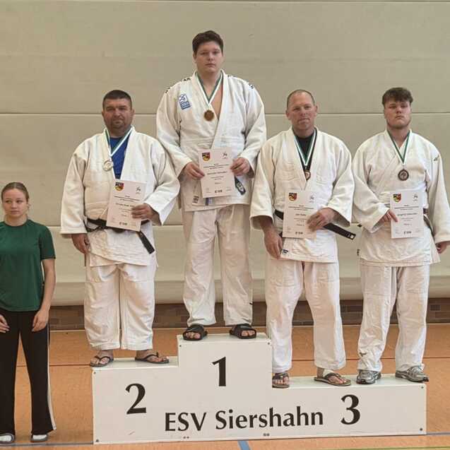 Drei Judokas in wei&szlig;en Anz&uuml;gen mit Medaillen stehen auf dem Siegertreppchen, w&auml;hrend zwei Zuschauer daneben stehen.